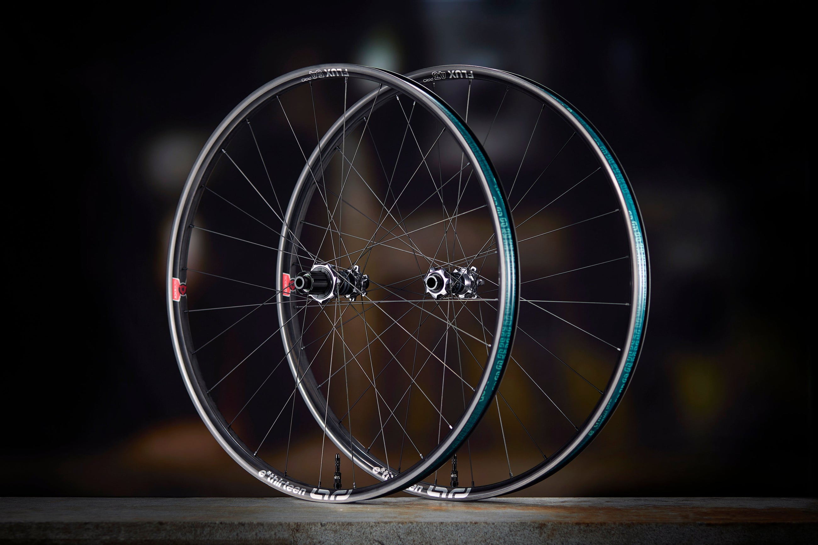 A Sidekick Flux front and rear wheel standing side by side on a dark floor, Flux rims with the teal rim-bed decal and Sidekick hubs.